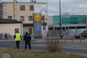 Widoczny policjant i strażnik miejski pieszo patrolujący ulicę. W dali widoczna kobieta prowadząca rower.