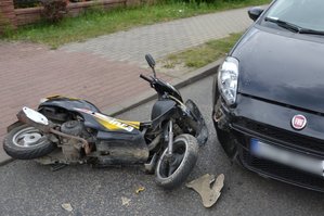 Widoczny roztrzaskany motorower, leżacy na jezdni przed samochodem osobowym, który jest częsciowo widoczny na zdjęciu.