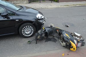 Widoczny roztrzaskany motorower, leżacy na jezdni przed samochodem osobowym, który jest częsciowo widoczny na zdjęciu.