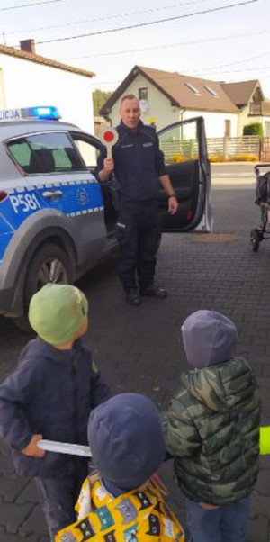 Dzielnicowy stojąc przy radiowozie pokazuje dzieciom lizak, którym policjanci posługują się kierując ruchem drogowym.