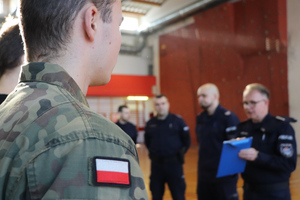 Na zdjęciu widoczna naszywka w postaci polskiej flagi, umieszczona na rękawie munduru zawodnika. W tle widoczni umundurowani policjanci.